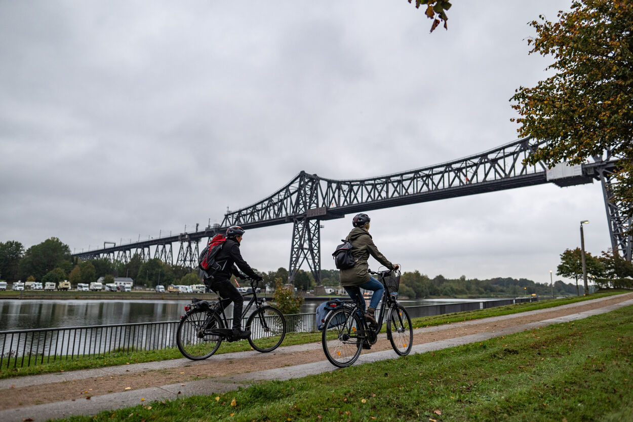 Zwei Radfahrende fahren entlang des Nord-Ostsee-Kanals zu der Eisenbahnhochbrücke in Rendsburg.