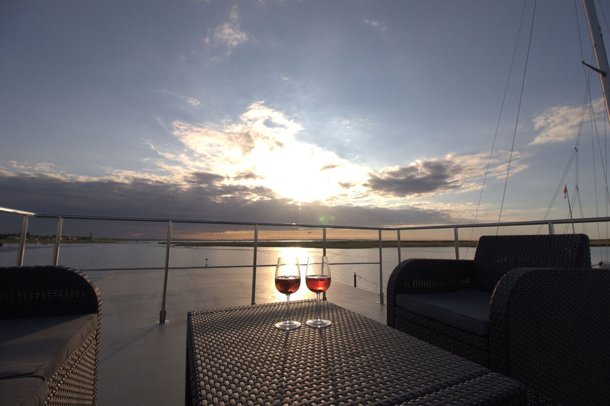 Terrasse mit Stühlen und einem Tisch auf einem Hausboot. Auf dem Tisch stehen zwei gefüllte Weingläser.